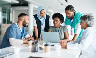 Diverse healthcare professionals collaborating around a laptop during a meeting, demonstrating teamwork and leadership in healthcare administration.